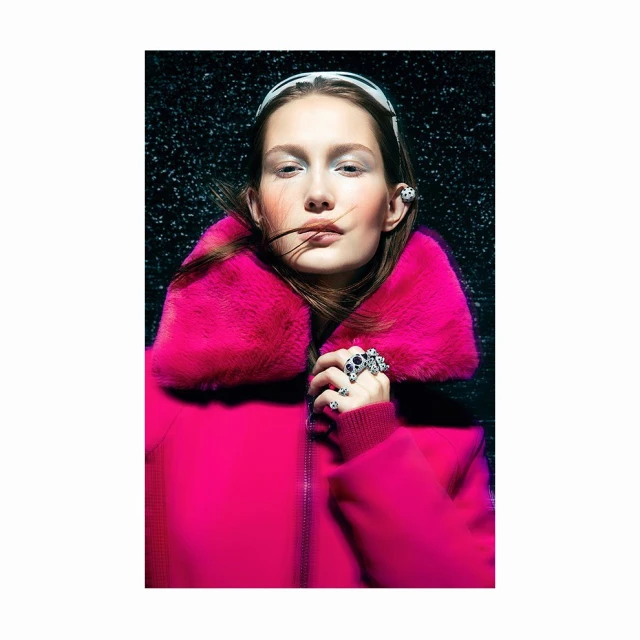 Fashion portrait of a woman wearing a pink coat and jewelry, standing against a black backdrop with water droplets.