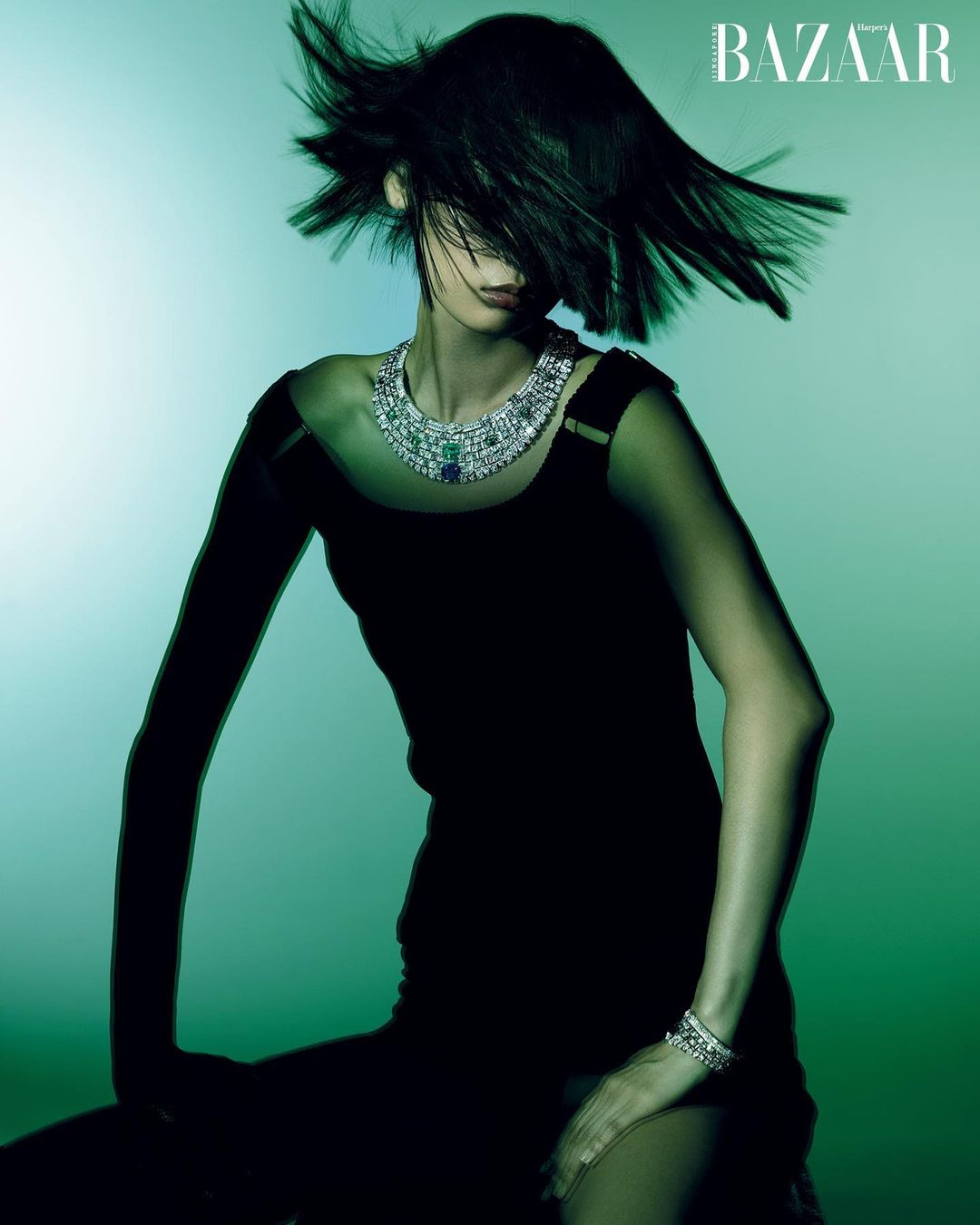 Fashion shot of woman in black dress wearing jewel-encrusted necklace and bracelet, shot in a studio with a green and white background.