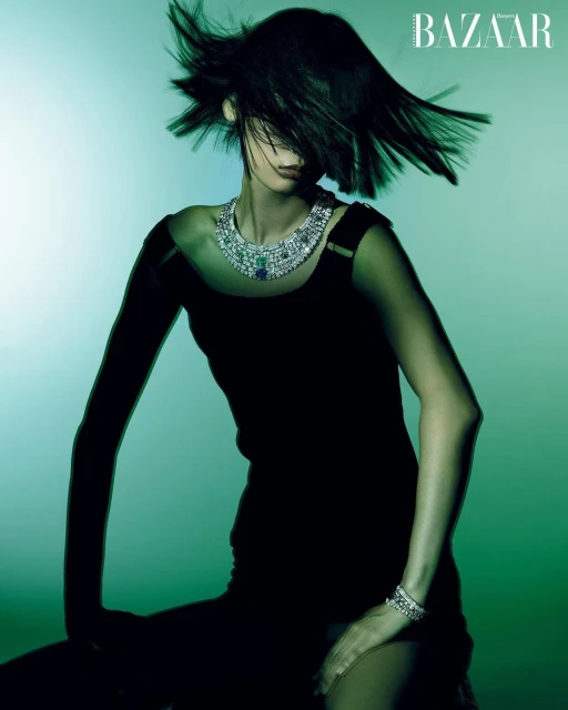 Fashion shot of woman in black dress wearing jewel-encrusted necklace and bracelet, shot in a studio with a green and white background.