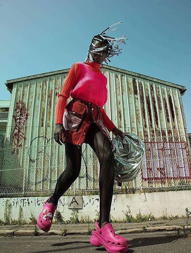 An adult woman poses on a street in hot-pink platform Crocs, a short red skirt, and a pink top against a corrugated metal wall.