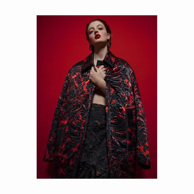 A woman poses in a patterned coat against a red backdrop, wearing dark bottoms and red lipstick in a studio fashion shot.