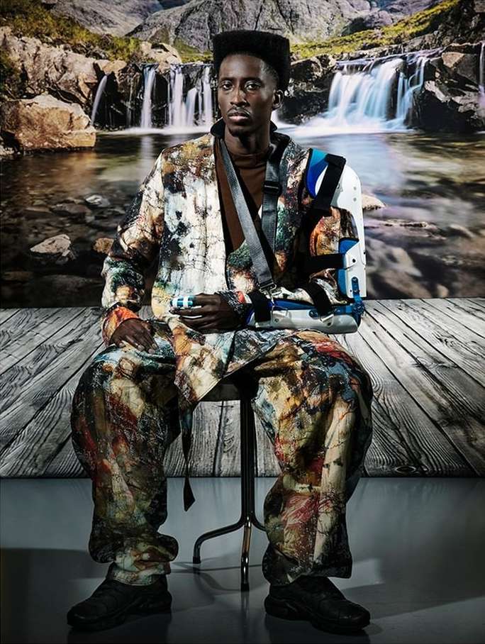 Adult man seated on a chair wearing a multicolored jacket and pants, a brown shirt, and an arm brace with a waterfall background.