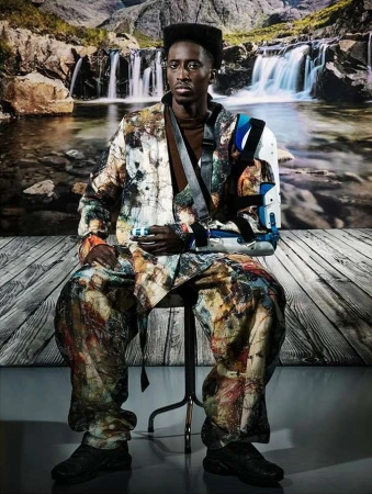 Adult man seated on a chair wearing a multicolored jacket and pants, a brown shirt, and an arm brace with a waterfall background.