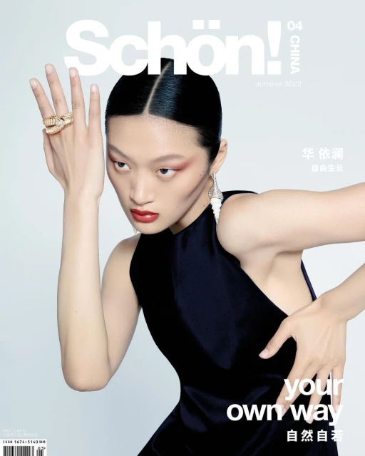 Magazine cover of a woman with slicked-back hair, red eye makeup, and dark dress posing with hand jewelry in a photo studio.