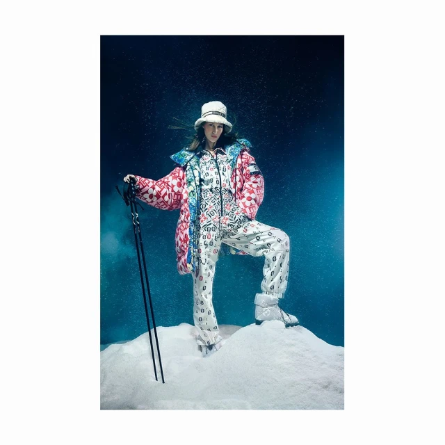 Woman in a patterned ski suit and hat posing on a snowy surface with ski poles, set against a dark blue backdrop for a fashion editorial.