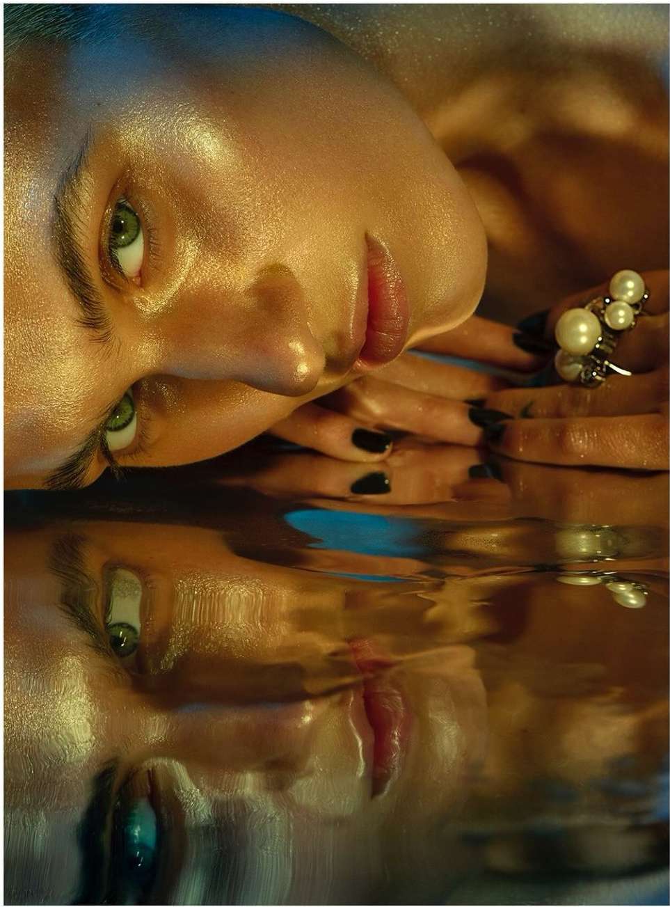 Close-up studio shot of a reflective portrait of a woman with green eyes, pearl ring, and black manicure in a blue and gold lighting.