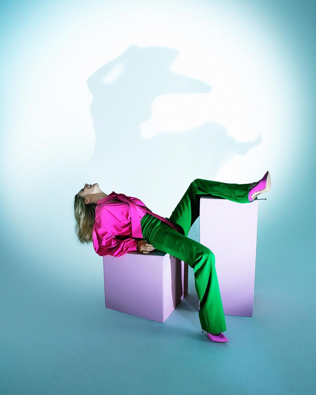 Fashion model reclines on two pink blocks in a photo studio wearing bright pink satin blouse, green pants, and matching heels. 