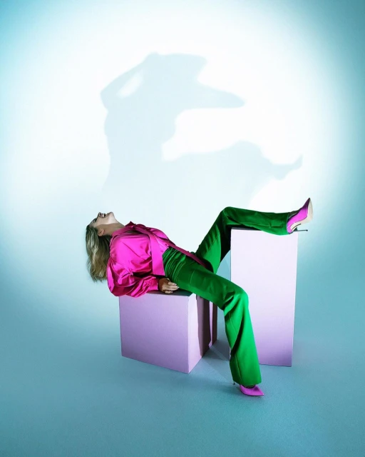 Fashion model reclines on two pink blocks in a photo studio wearing bright pink satin blouse, green pants, and matching heels. 
