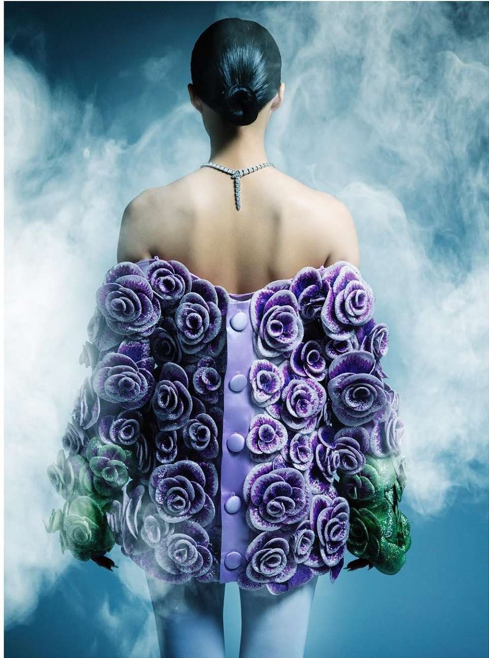 Back view of a person with dark hair, wearing a purple floral jacket, and a diamond necklace against a smoky blue background.