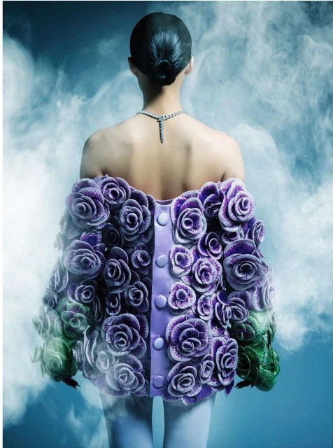 Back view of a person with dark hair, wearing a purple floral jacket, and a diamond necklace against a smoky blue background.