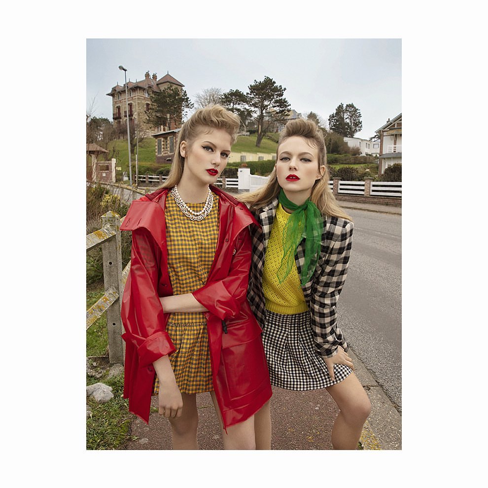Two women in retro fashion stand on a street near houses and trees, one in a red coat and the other in a plaid blazer with a green scarf.