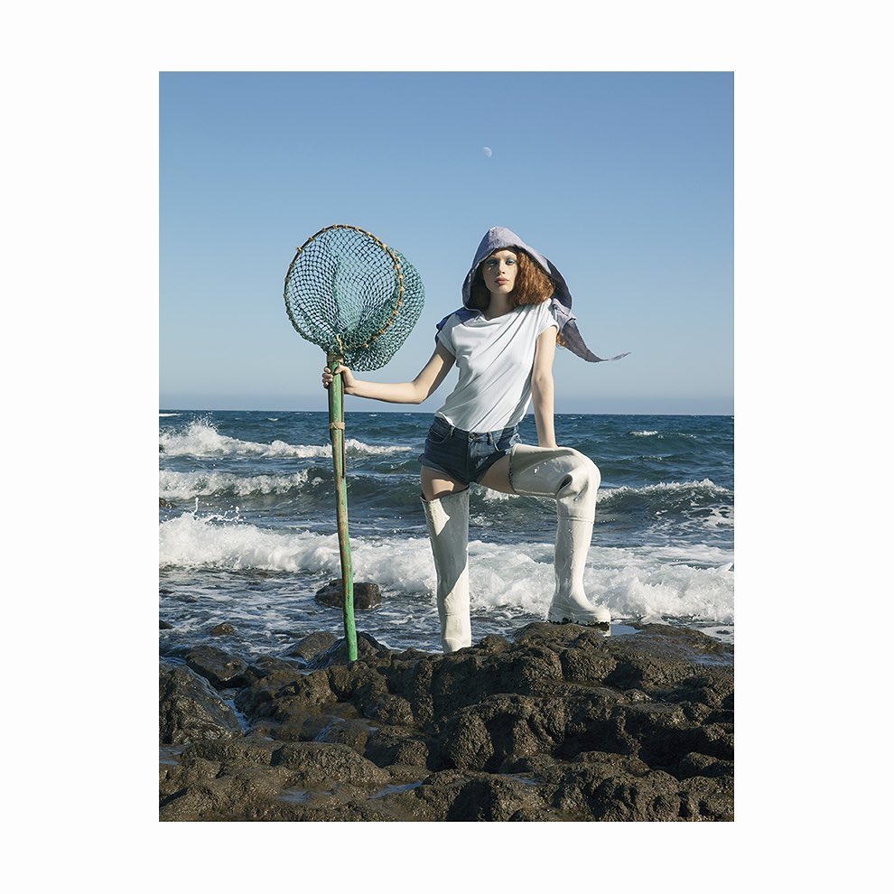 Fashionable young woman posing on rocks by ocean with fishing net. She is wearing a hooded shirt, shorts, high boots in outdoor setting.