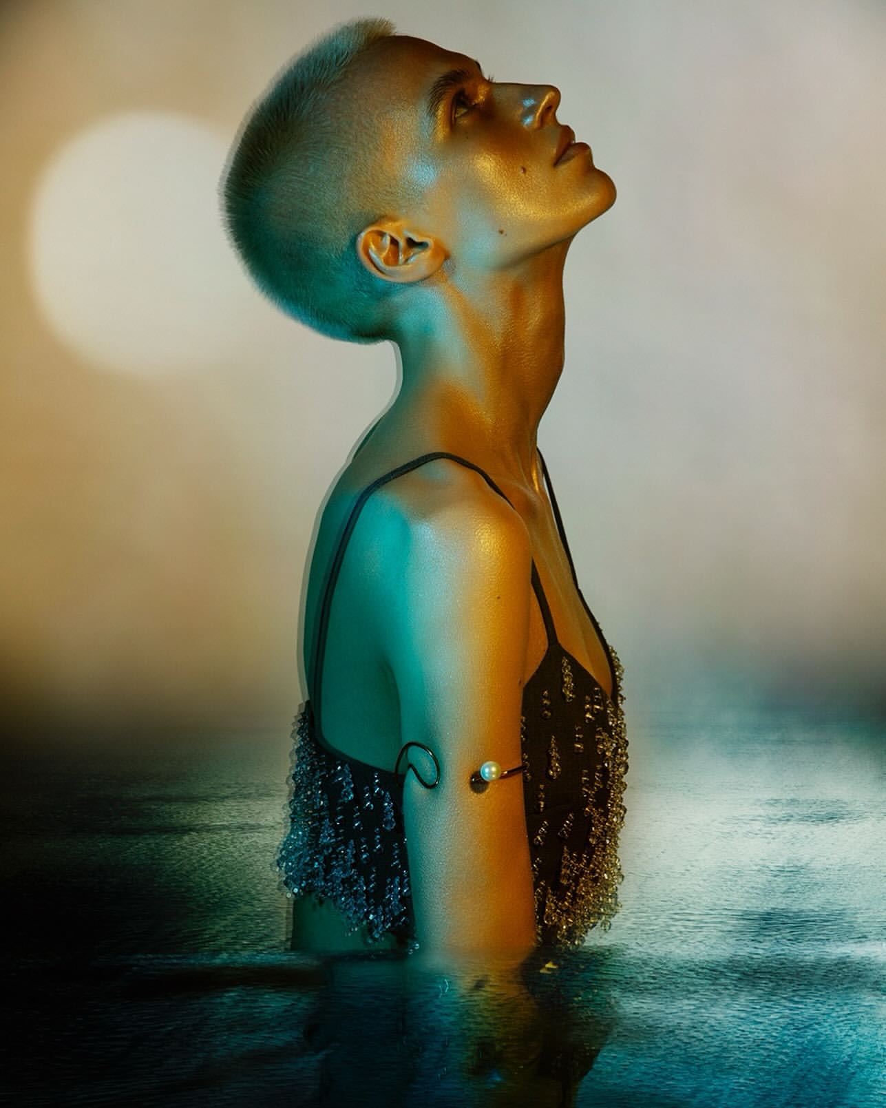 Side view of a person with a shaved head and metallic body paint in a black beaded top standing in shallow water with a moon background.