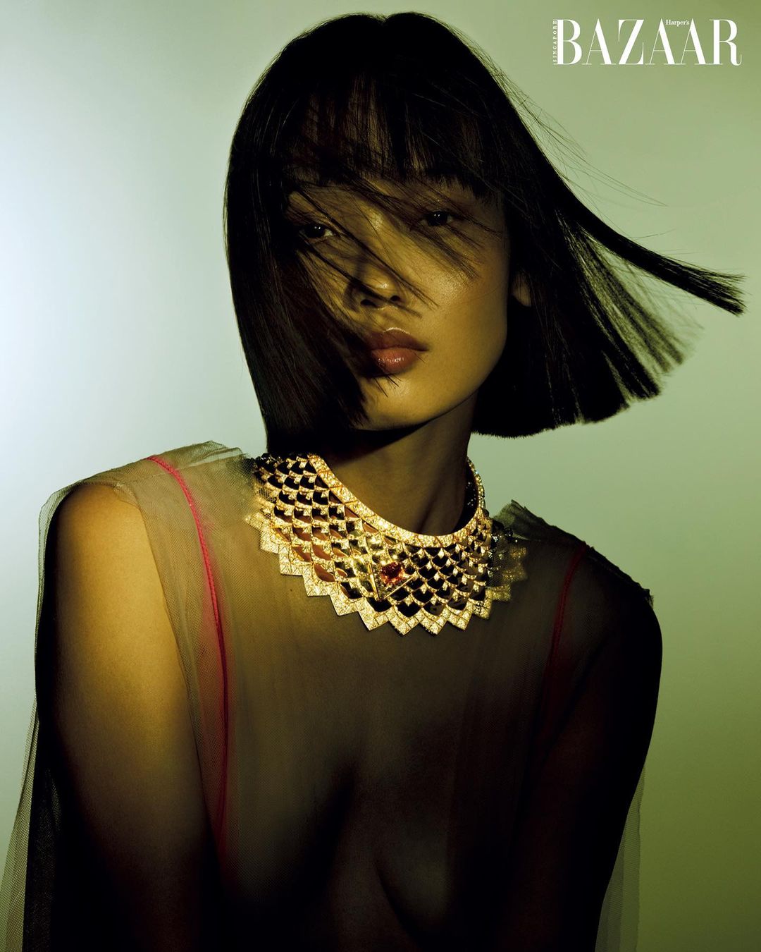 Elegant woman in a sheer top and a diamond necklace is featured in a studio shot with BAZAAR logo. The subject is looking towards the camera.