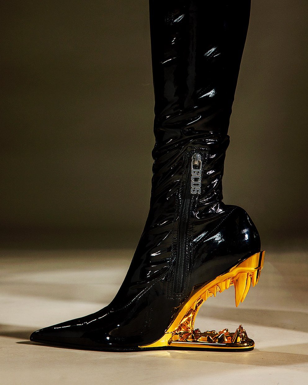 A black, high, knee-high, glossy patent-leather boot with a gold colored, open, jagged platform heel is displayed in a studio shot.