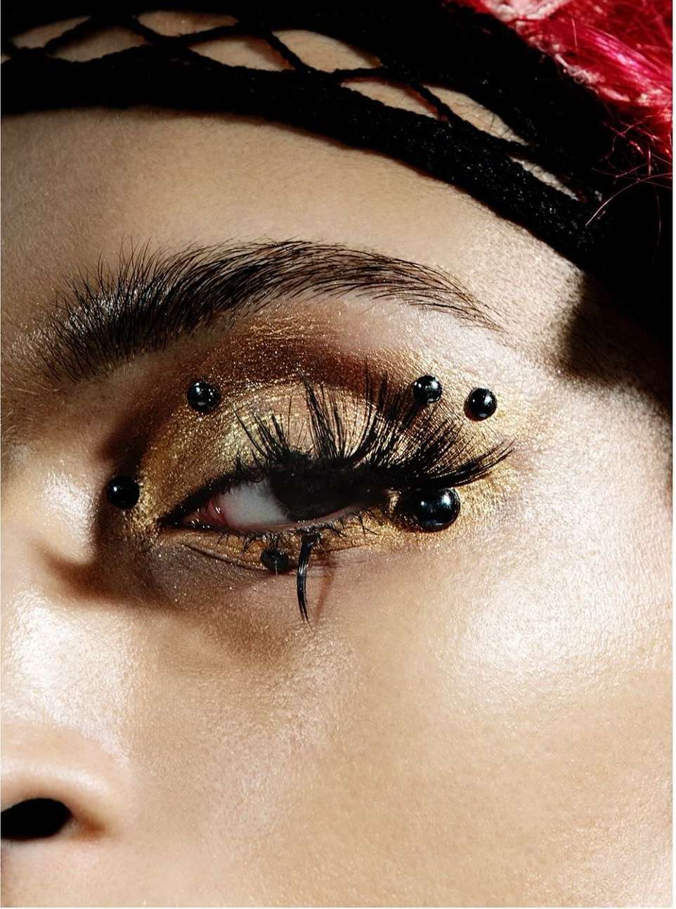 Close-up of an adult female eye with dramatic gold eye makeup, black eyelash extensions, and black decorative facial piercings.