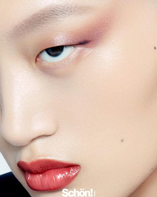 Close-up portrait of a young adult female with flawless skin, glossy lips, and pink eyeshadow posing in a beauty or fashion editorial.