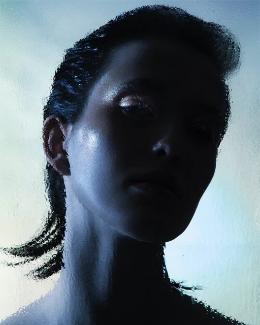 Close-up studio portrait of a woman with dark hair, lit with dramatic lighting and refracted through textured glass, mysterious and stylish.