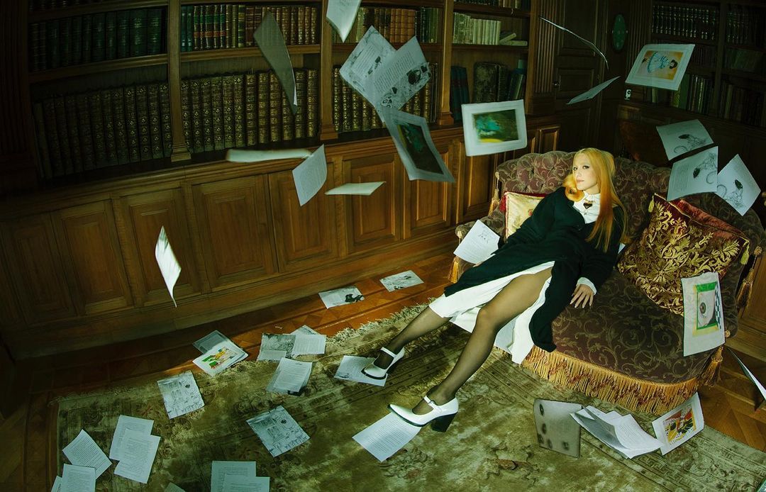 Woman reclines on a patterned sofa in a room with a floor-to-ceiling bookshelf as loose papers float around her and on the floor.