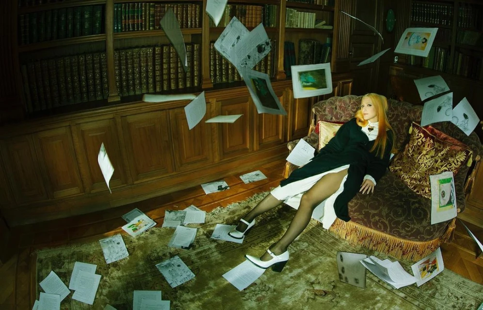 Woman reclines on a patterned sofa in a room with a floor-to-ceiling bookshelf as loose papers float around her and on the floor.
