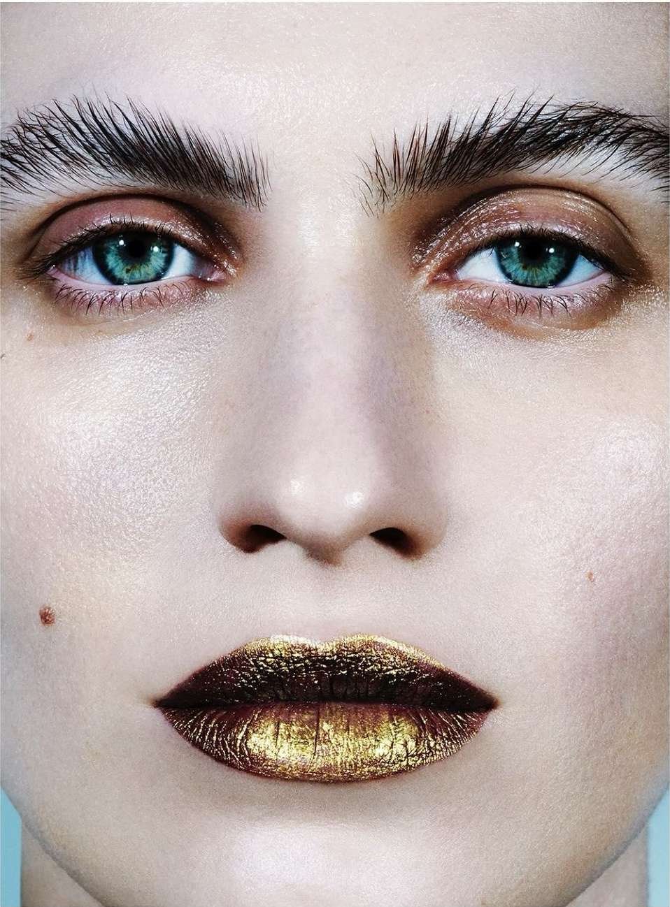 Extreme close-up of a model's face with bold makeup, featuring a focus on teal eyes, strong brow styling, and gold metallic lips.
