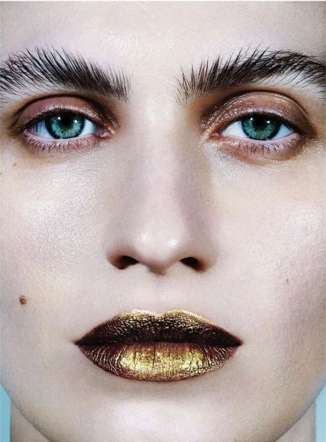 Extreme close-up of a model's face with bold makeup, featuring a focus on teal eyes, strong brow styling, and gold metallic lips.