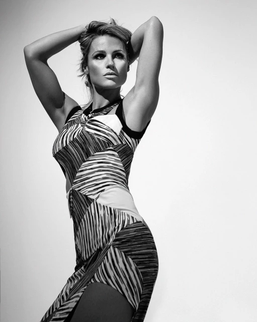 Black and white full body portrait of an adult woman with hands behind her head wearing a patterned sleeveless dress against a neutral backdrop.