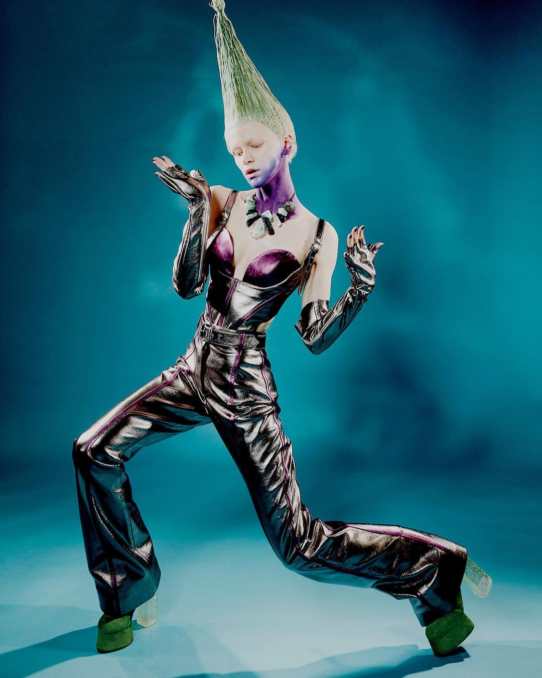 Woman with green cone-shaped hair poses in a silver and purple jumpsuit with matching gloves against a blue background in a studio shot.