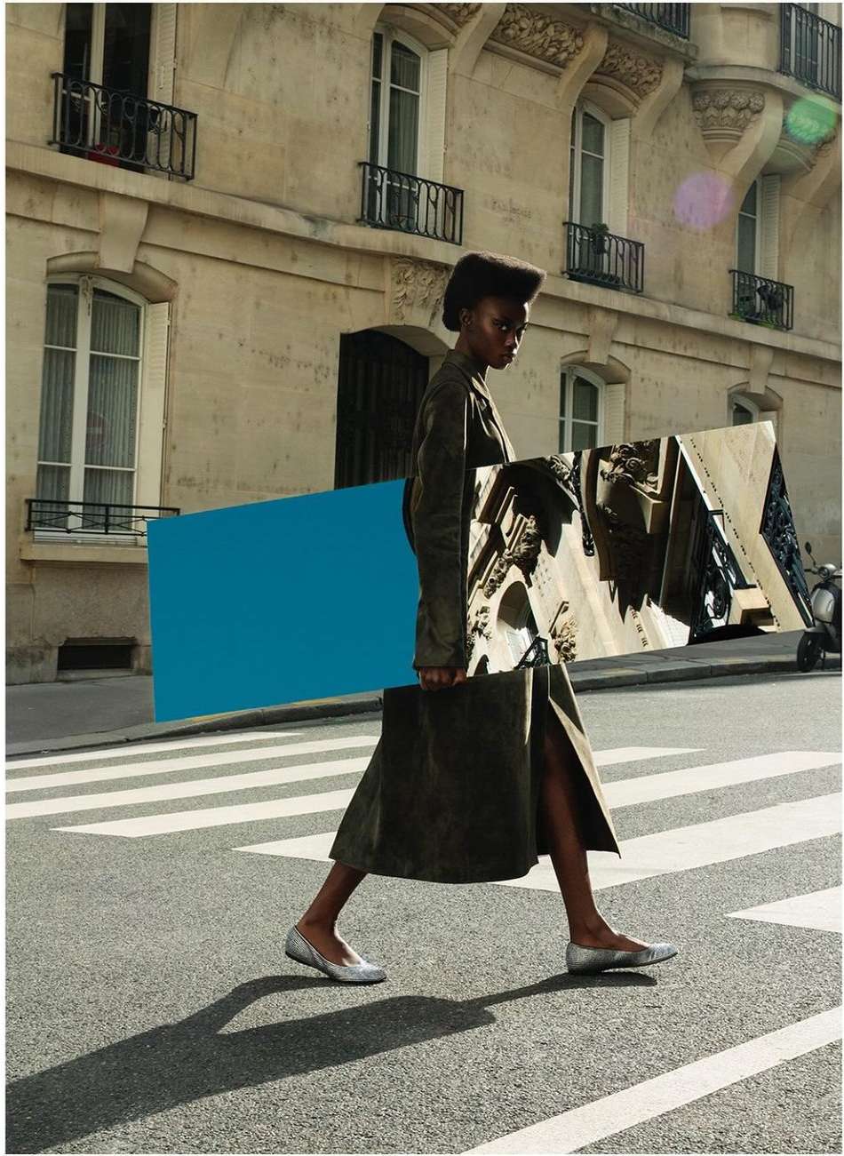 A woman walks across a crosswalk in an urban setting, carrying a blue and mirrored geometric shape, wearing a long coat and flats.