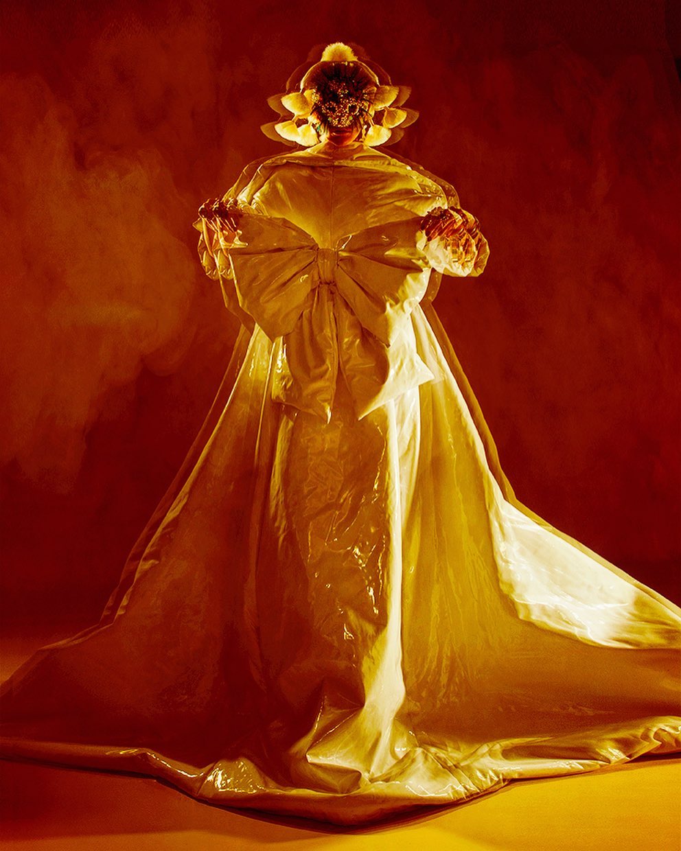 Person in yellow gown with large bow posing in photo studio with dramatic lighting and a red background creating a surreal fashion image.