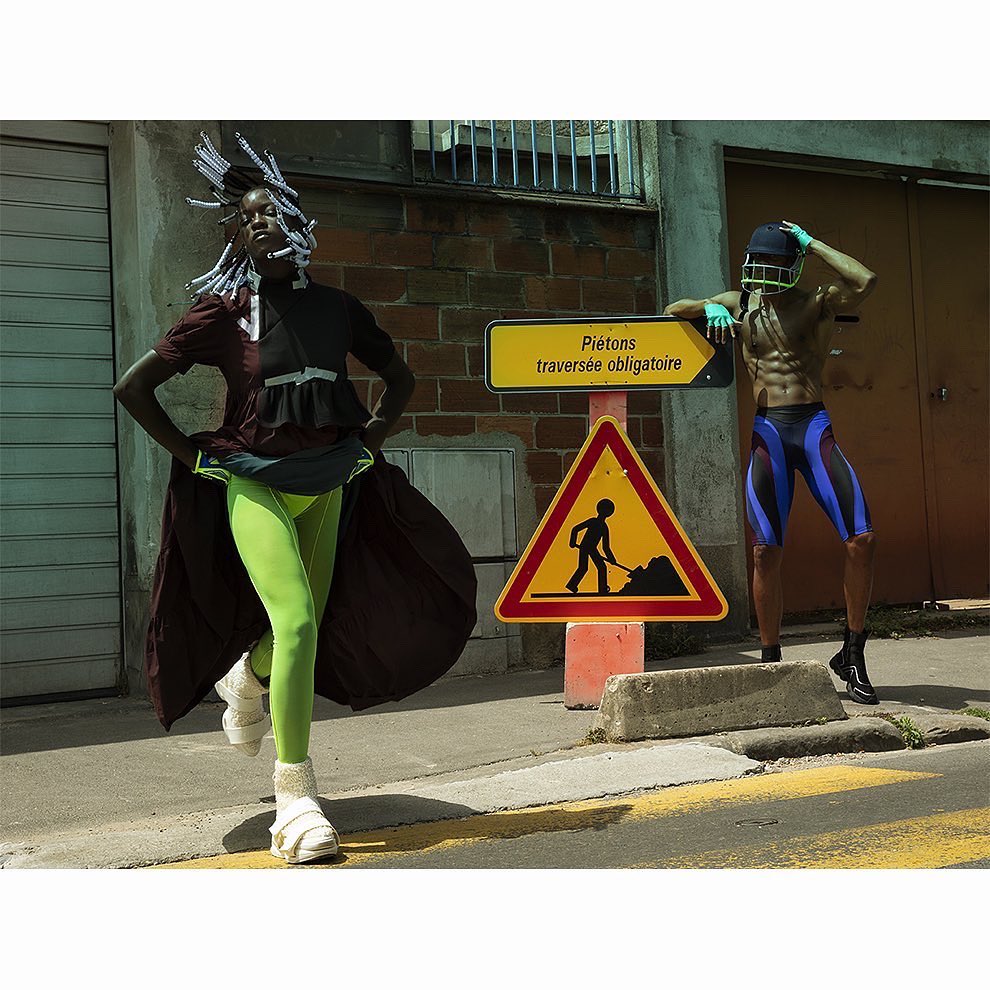 Two fashion models pose near a construction sign in athletic and eclectic attire, with one in neon green leggings and the other shirtless.