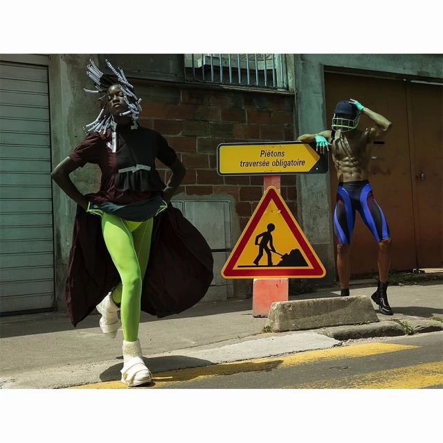 Two fashion models pose near a construction sign in athletic and eclectic attire, with one in neon green leggings and the other shirtless.