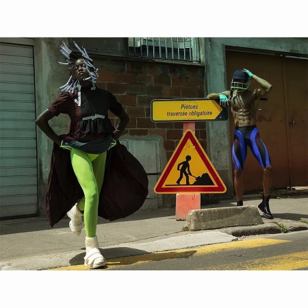 Two fashion models pose near a construction sign in athletic and eclectic attire, with one in neon green leggings and the other shirtless.