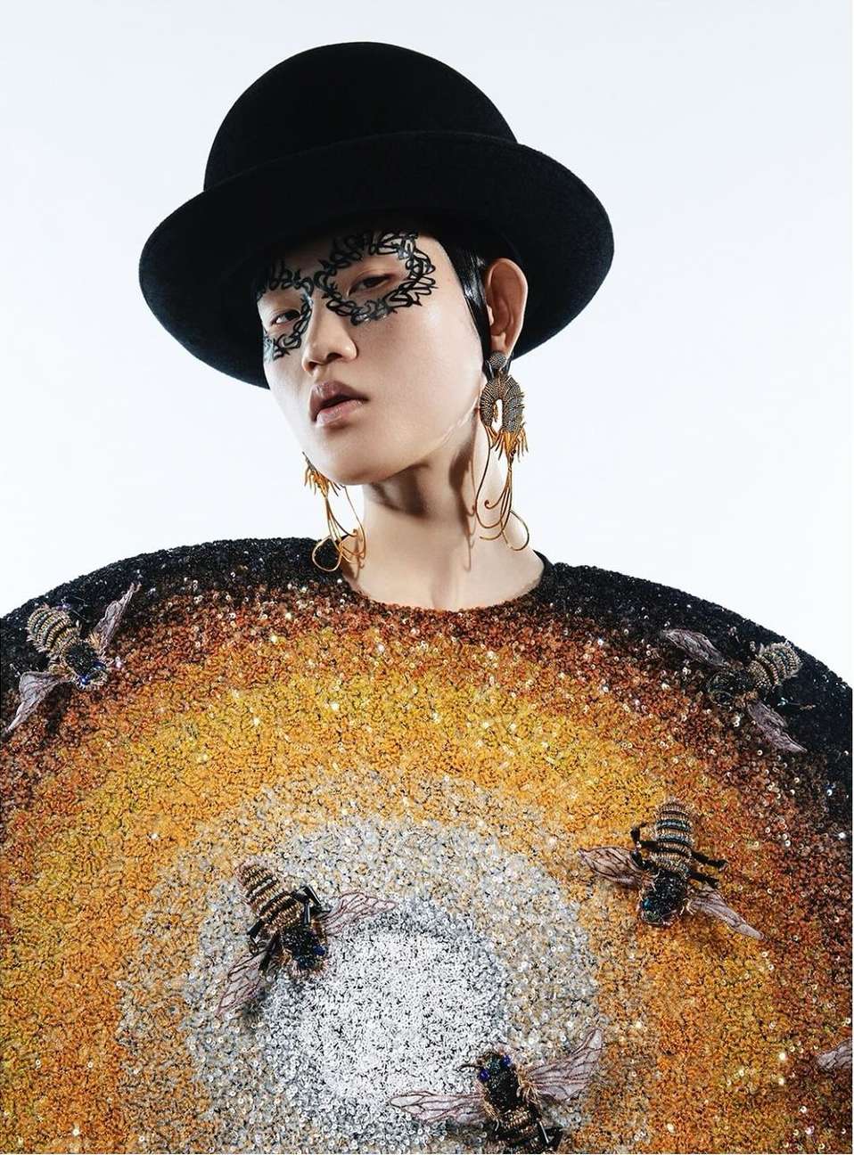 An Asian model showcases a creative fashion look with an embellished top, striking face makeup, dangling earrings, and a stylish hat.