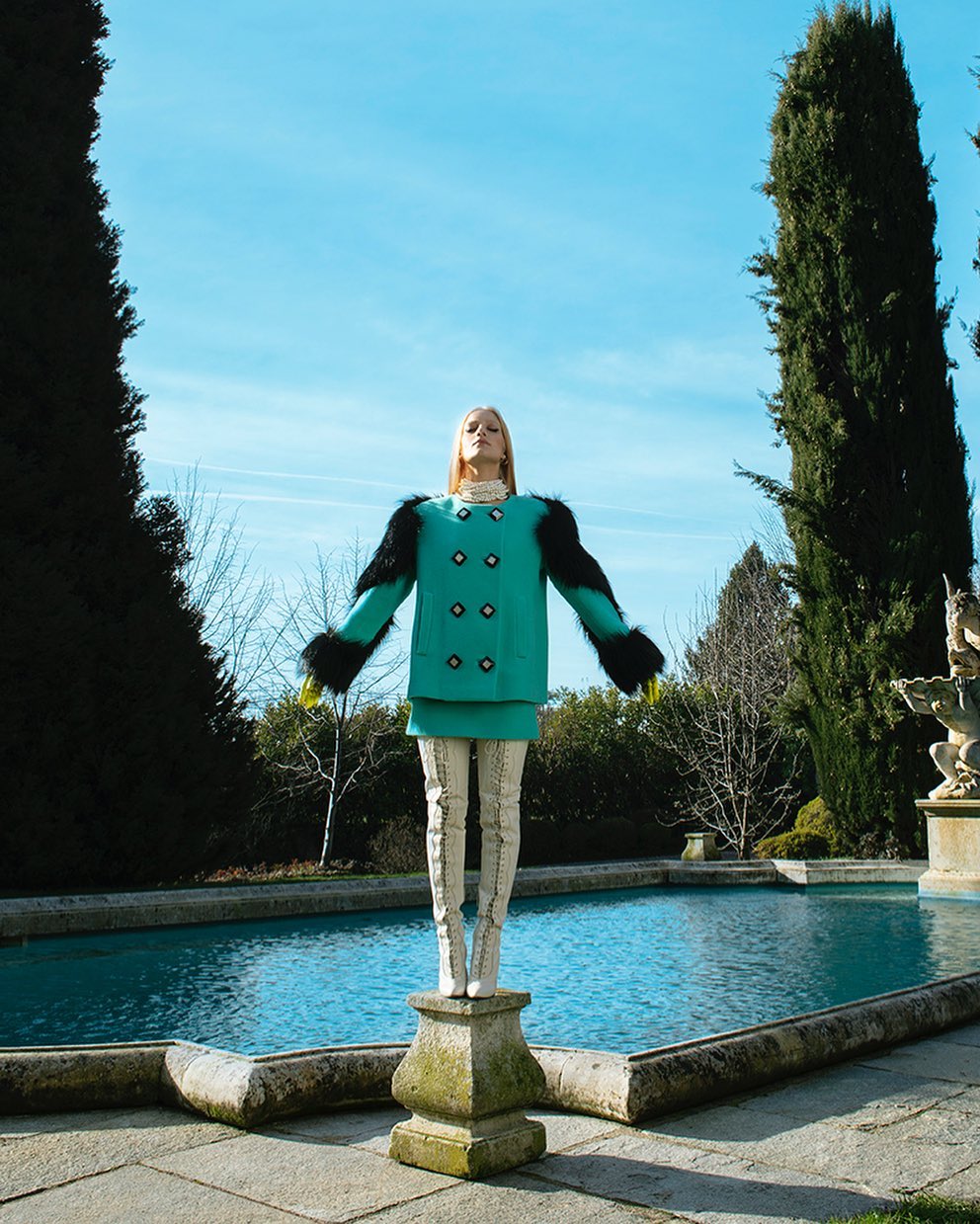 Woman in blue coat with fur trim posing on a stone pedestal near a turquoise pool outdoors on a sunny day with a clear blue sky.
