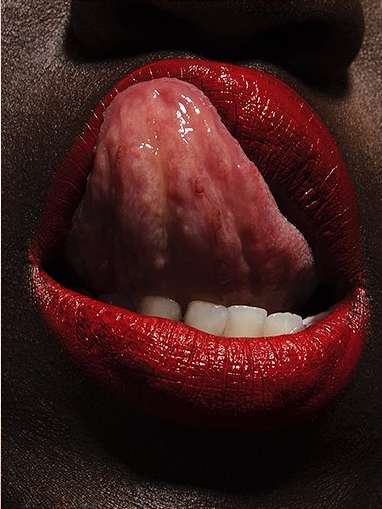 Close-up studio shot of a dark-skinned woman with bright red lipstick, bared teeth, and tongue extended out of her mouth.