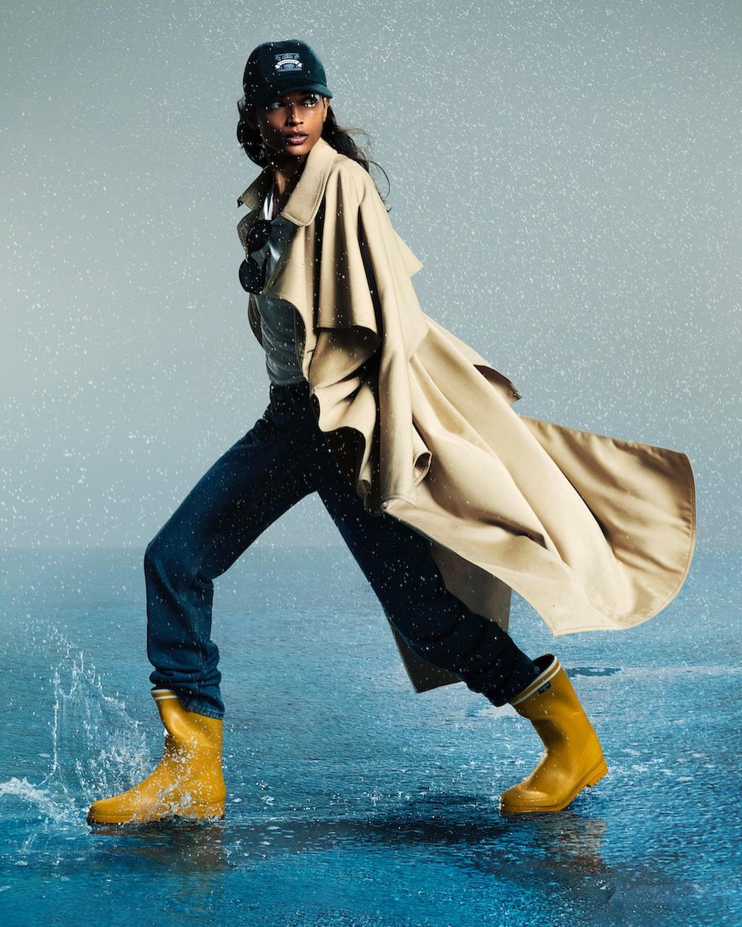 A woman in a trench coat and yellow boots walks in the rain. Her outfit includes dark pants and a baseball cap. She is walking through puddles.