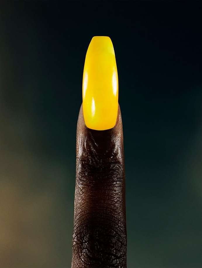 Close-up studio shot showcases an adult's brown-skinned finger with a striking yellow acrylic nail, set against a blurred backdrop.