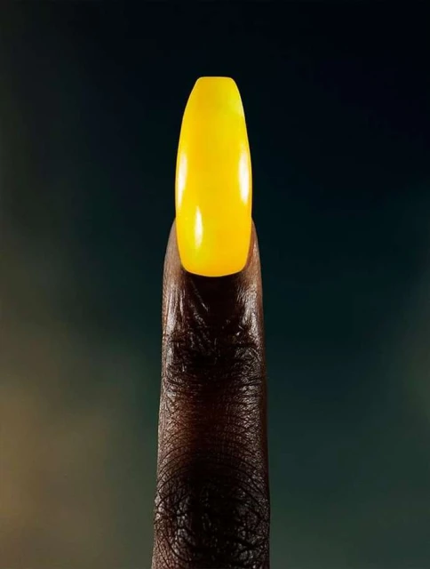 Close-up studio shot showcases an adult's brown-skinned finger with a striking yellow acrylic nail, set against a blurred backdrop.