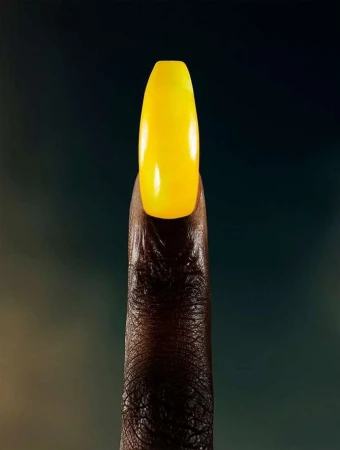 Close-up studio shot showcases an adult's brown-skinned finger with a striking yellow acrylic nail, set against a blurred backdrop.