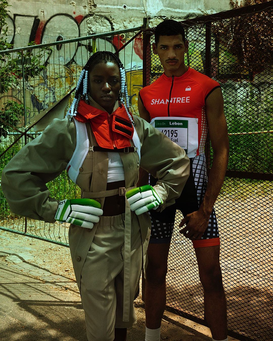 Two people pose in athletic wear against a graffiti wall. One is in an olive-green ensemble, the other in red and black athletic wear.