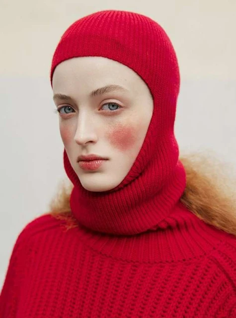 Close-up of a fair-skinned woman with blue eyes wearing a red knit balaclava and turtleneck sweater, red blush on her cheeks.