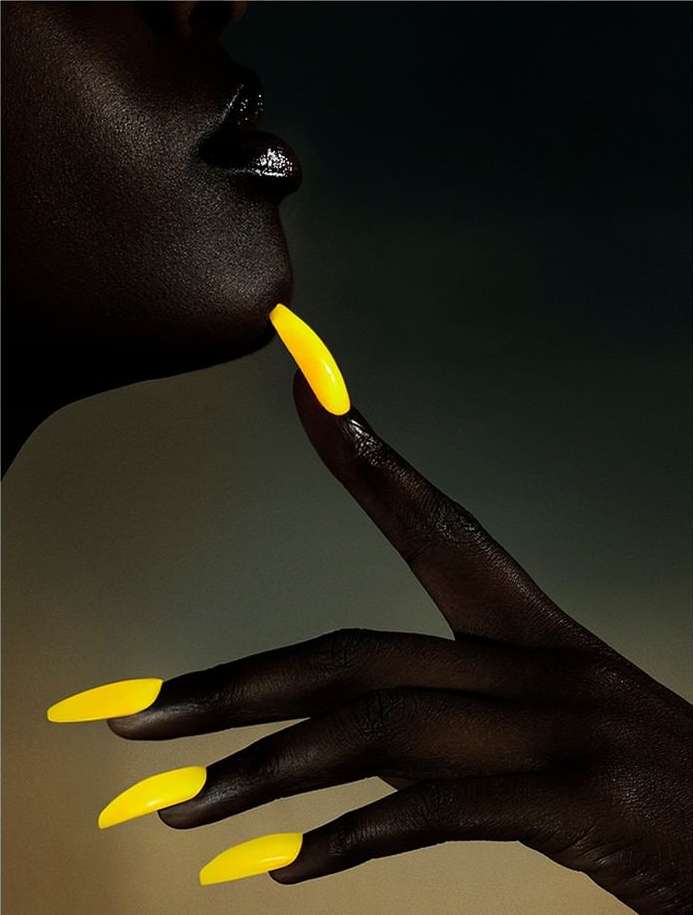 Close-up of a Black woman's face and hand with bright yellow, stiletto-shaped nails, illuminated against a dark gradient background.