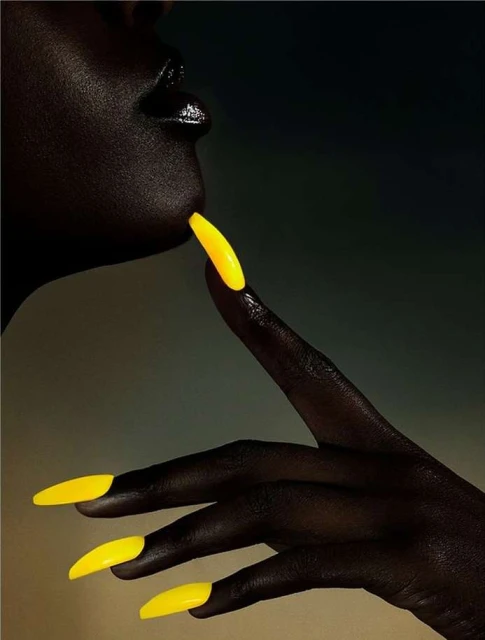 Close-up of a Black woman's face and hand with bright yellow, stiletto-shaped nails, illuminated against a dark gradient background.