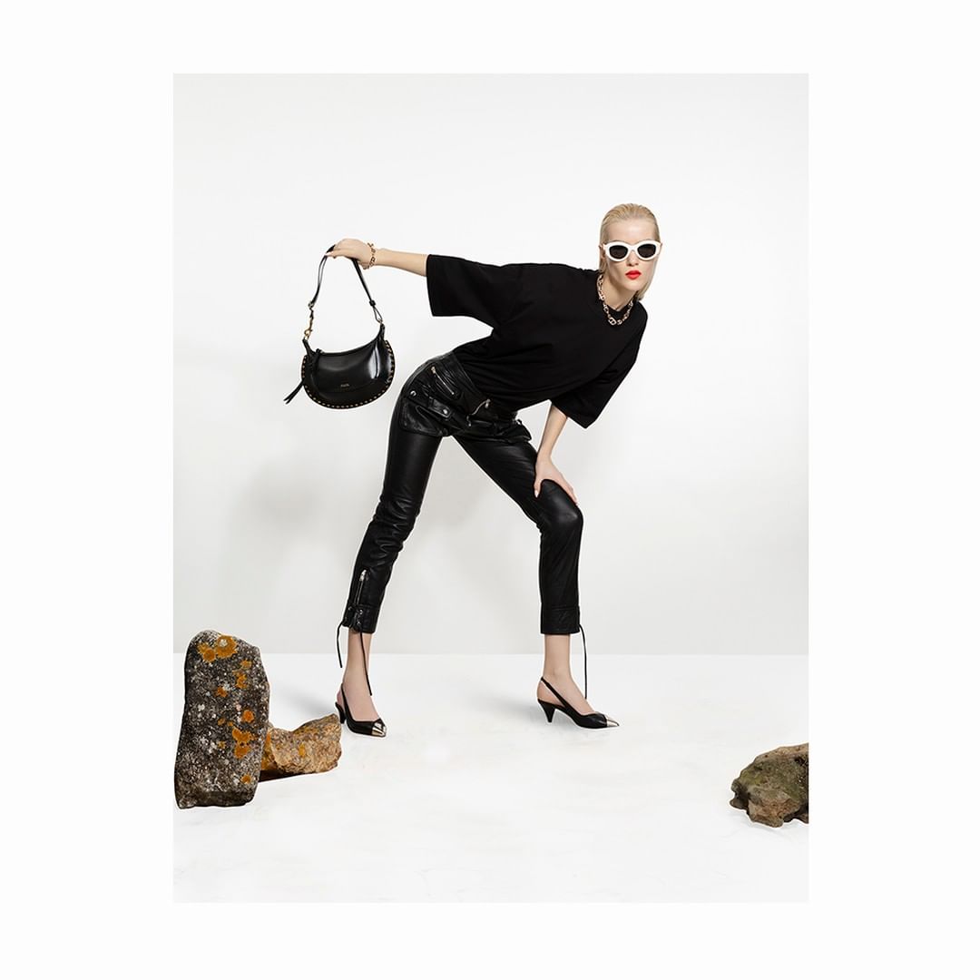 Model in black t-shirt and leather trousers holding a handbag against white studio backdrop with rocks