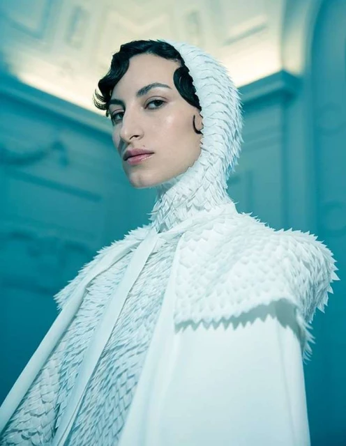 Portrait of a woman wearing a white hooded dress with textured detail, posing in an indoor setting with light-blue toned walls.