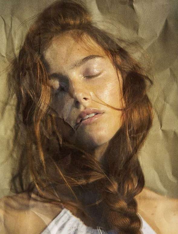 Close-up portrait of a red-haired woman with fair skin and closed eyes, lying against a neutral beige background in studio lighting.