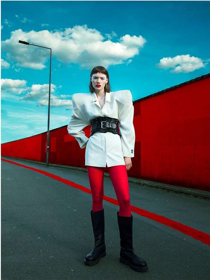 Woman in white blazer dress, black belt, red tights, and black boots stands on an asphalt road against red wall and cloudy blue sky.
