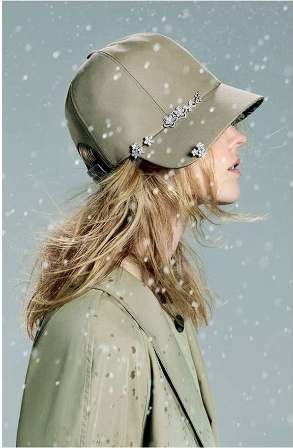 A woman with fair hair is seen in a close-up shot wearing a khaki cap decorated with silver embellishments, and a matching trench coat.