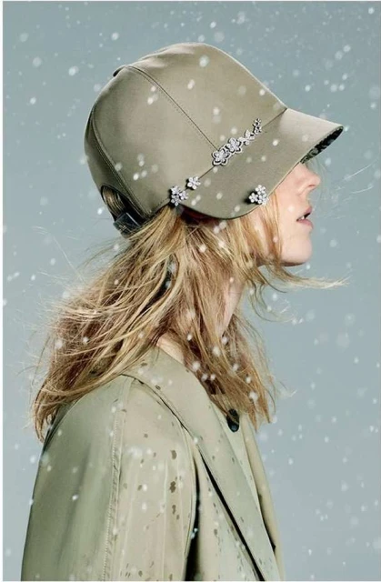 A woman with fair hair is seen in a close-up shot wearing a khaki cap decorated with silver embellishments, and a matching trench coat.